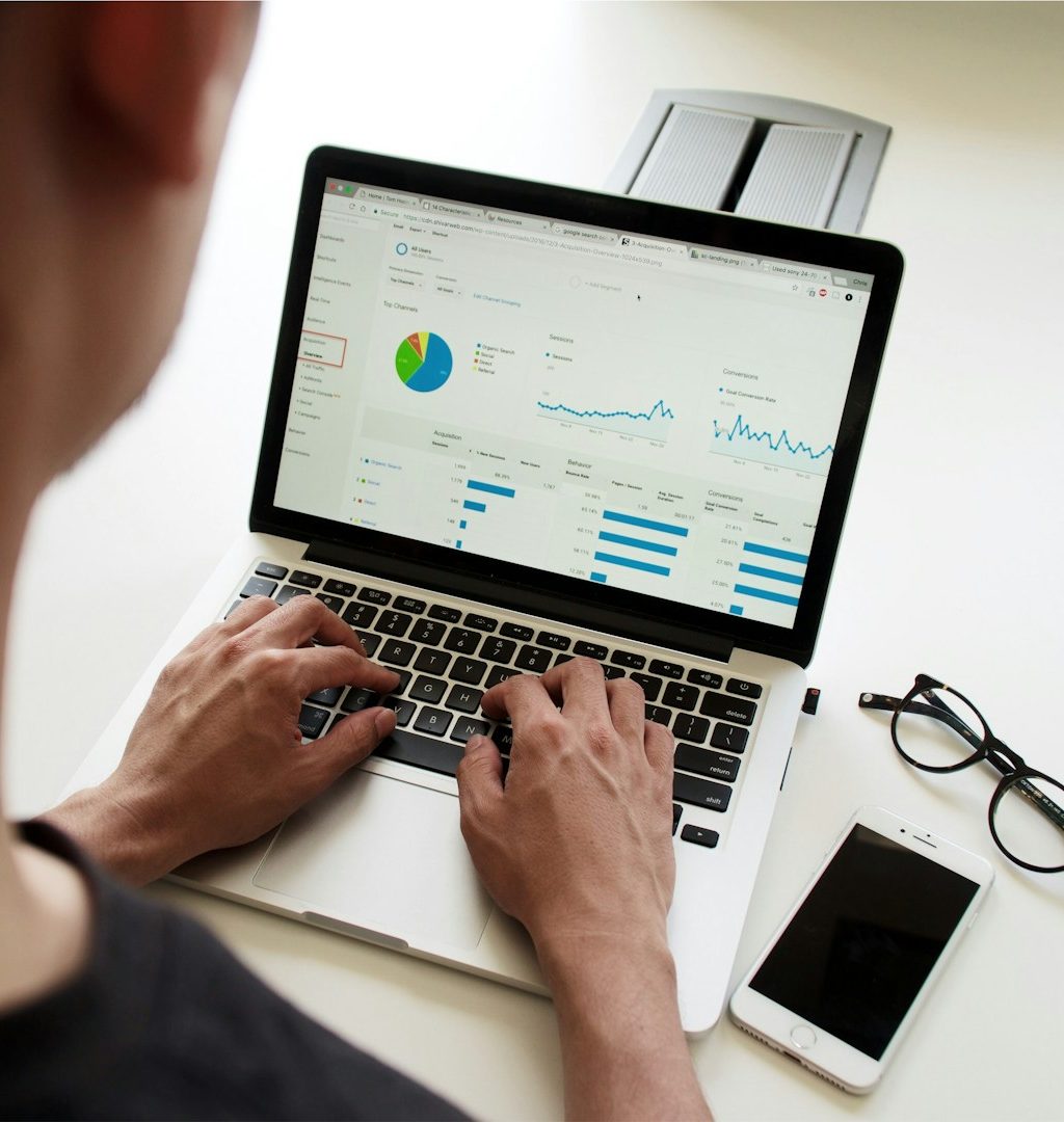Person arbeitet an einem Laptop mit Datenanalysen, daneben ein Smartphone und eine Brille.
